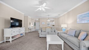 Una sala de estar con televisor, muebles blancos, ventilador de techo y un cuadro en la pared.