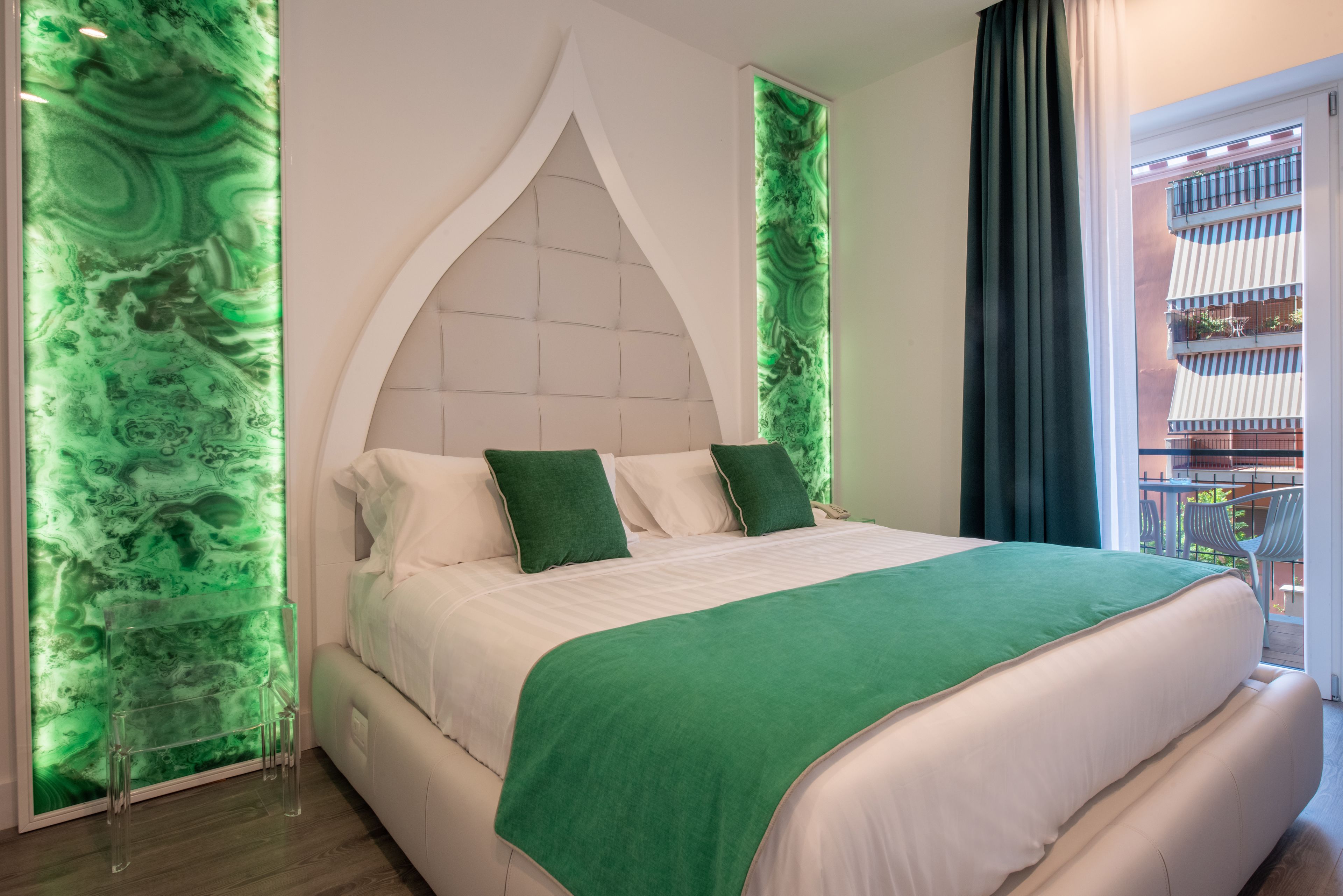 A modern bedroom with a large bed, green accents, and a view of a balcony with outdoor furniture.