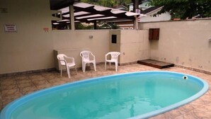 An outdoor swimming pool with a tiled floor, surrounded by a low wall and a covered seating area with plastic chairs.
