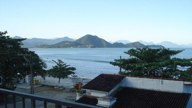 A coastal view with a balcony overlooking the sea, a tiled roof, and distant mountains.