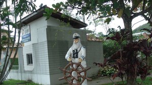 A statue of a bearded man holding a ship's wheel, standing on a green platform in front of a white building with a sign that reads "Toninhas."