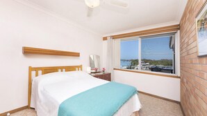 Une chambre à coucher avec un lit, une fenêtre donnant sur les bateaux et l’eau, une tête de lit en bois et un ventilateur de plafond.