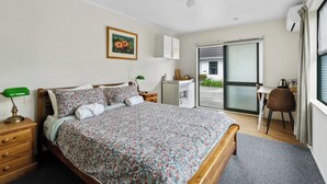 A bedroom with a bed, a desk, a chair, a wardrobe, and a sliding glass door leading to an outdoor area. - Atarau Grove  Studio (Paraparaumu)