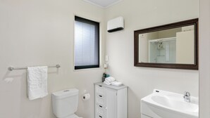 A bathroom with a toilet, a cabinet with drawers, a sink, a mirror, and a window with blinds. - Atarau Grove  Studio (Paraparaumu)