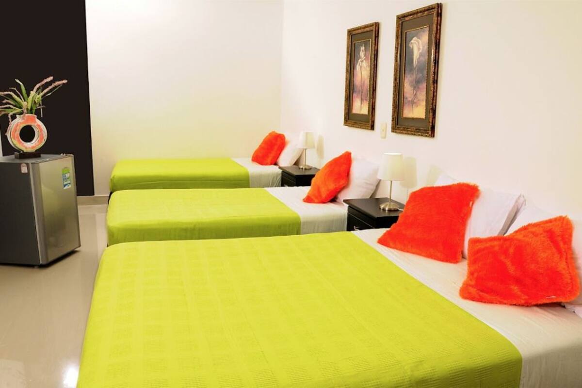A hotel room with three beds, each with a green bedspread and orange pillows. There are two framed pictures on the wall, a small table with a lamp, and a refrigerator on the left.