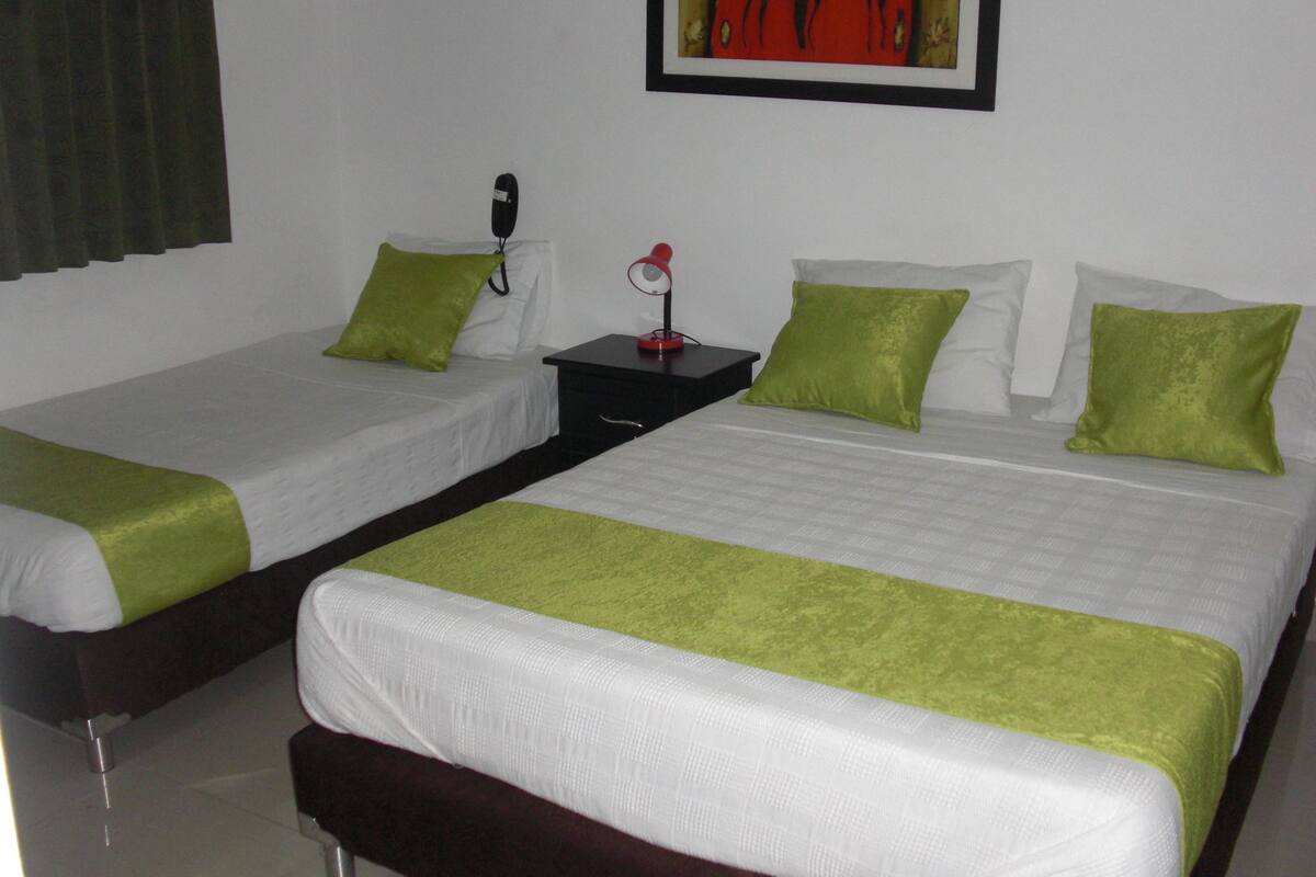 A hotel room with two single beds, each with a green pillow and white bedding. There is a nightstand with a telephone and a red lamp between the beds.