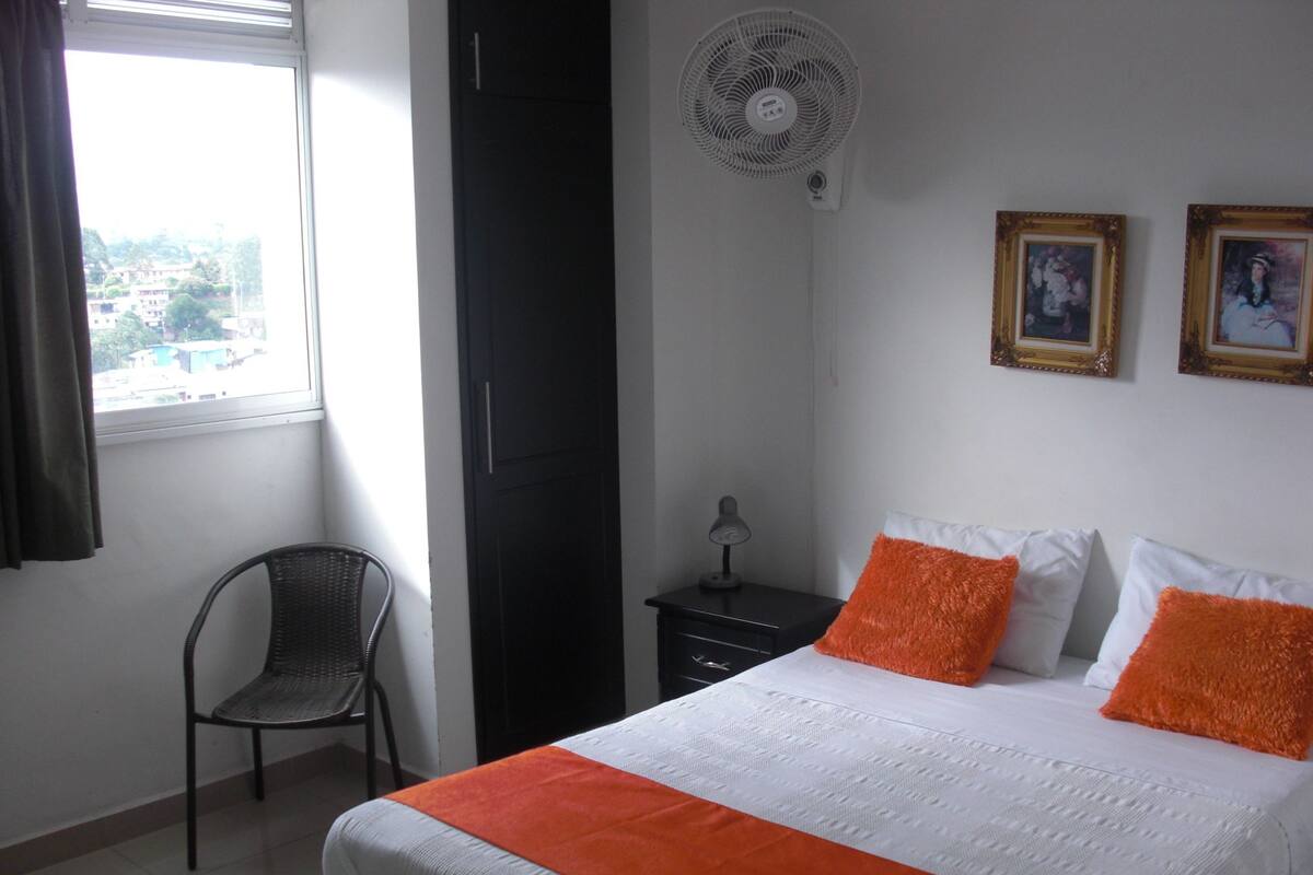 A bedroom with a bed, a chair, a nightstand, a wall-mounted fan, and two framed pictures on the wall.