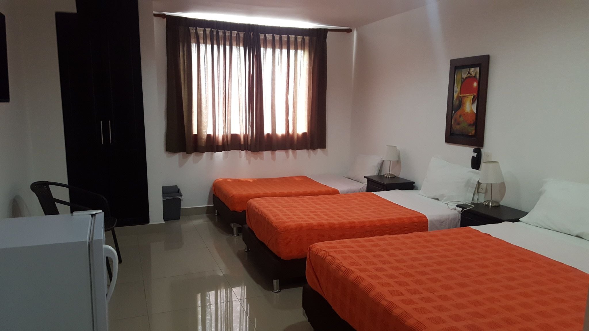 A hotel room with two beds, a desk, a chair, a window with curtains, a painting on the wall, and a trash bin.