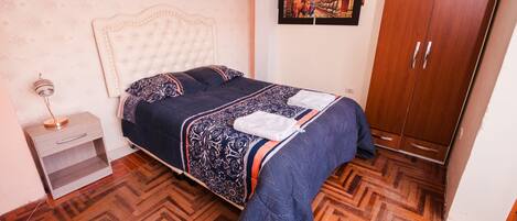 A bedroom with a bed, a wooden wardrobe, a nightstand, and a wall-mounted TV.