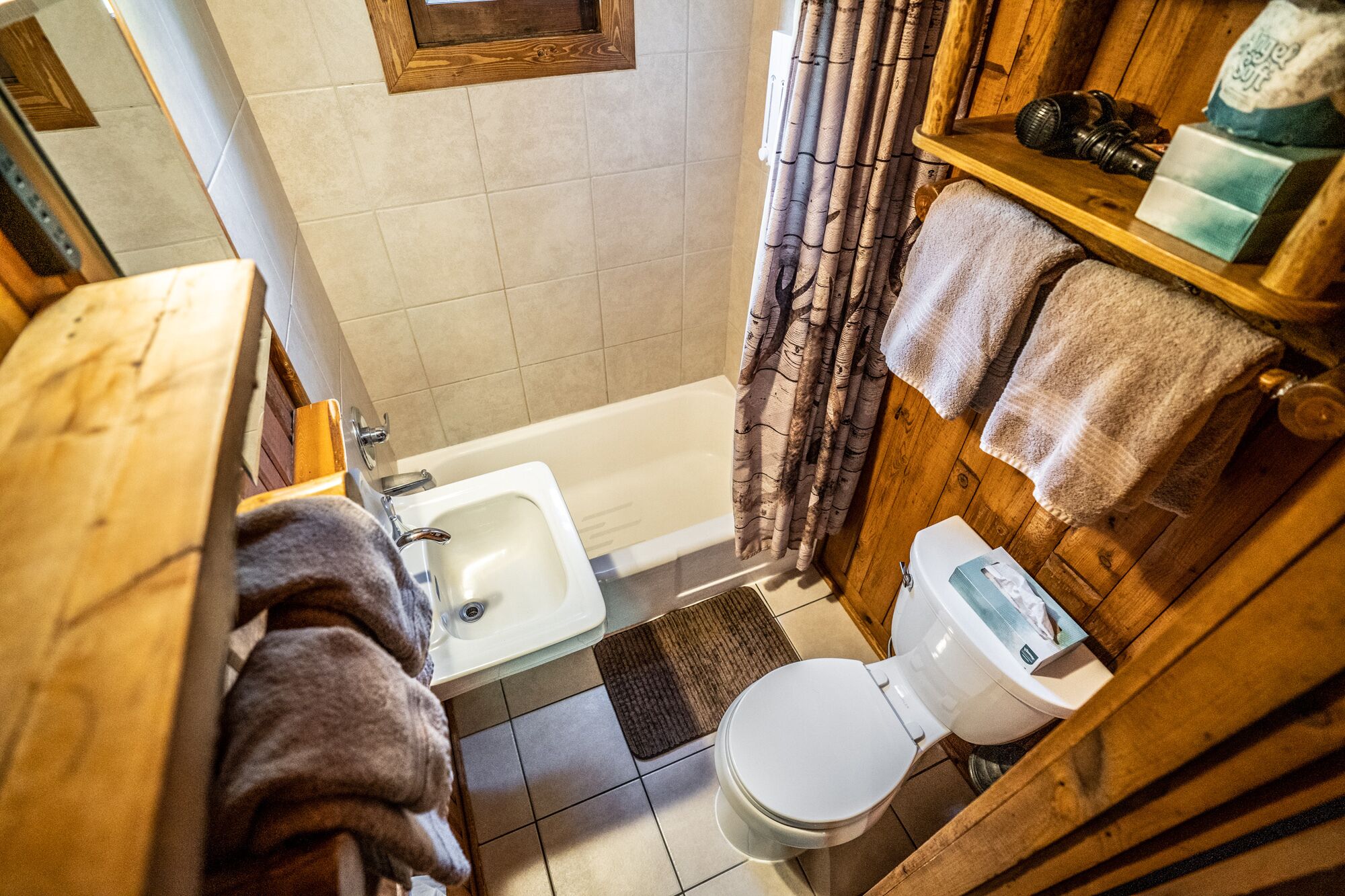 Baño compacto con lavabo, inodoro y bañera. Un estante de madera sostiene toallas y una cámara.