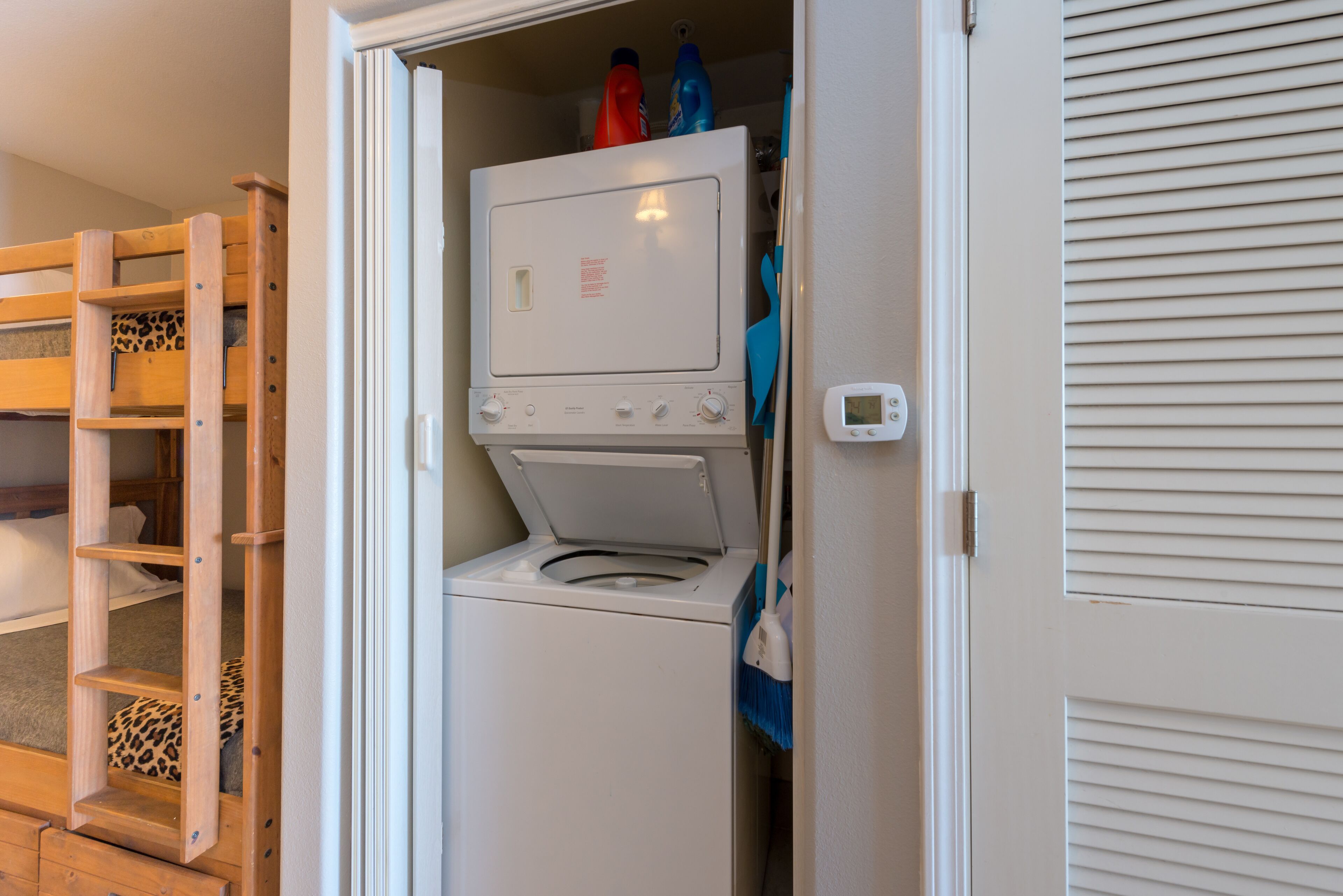 A compact laundry room with a front-loading washer and dryer, a wooden shelf unit, and a door with a temperature control device.