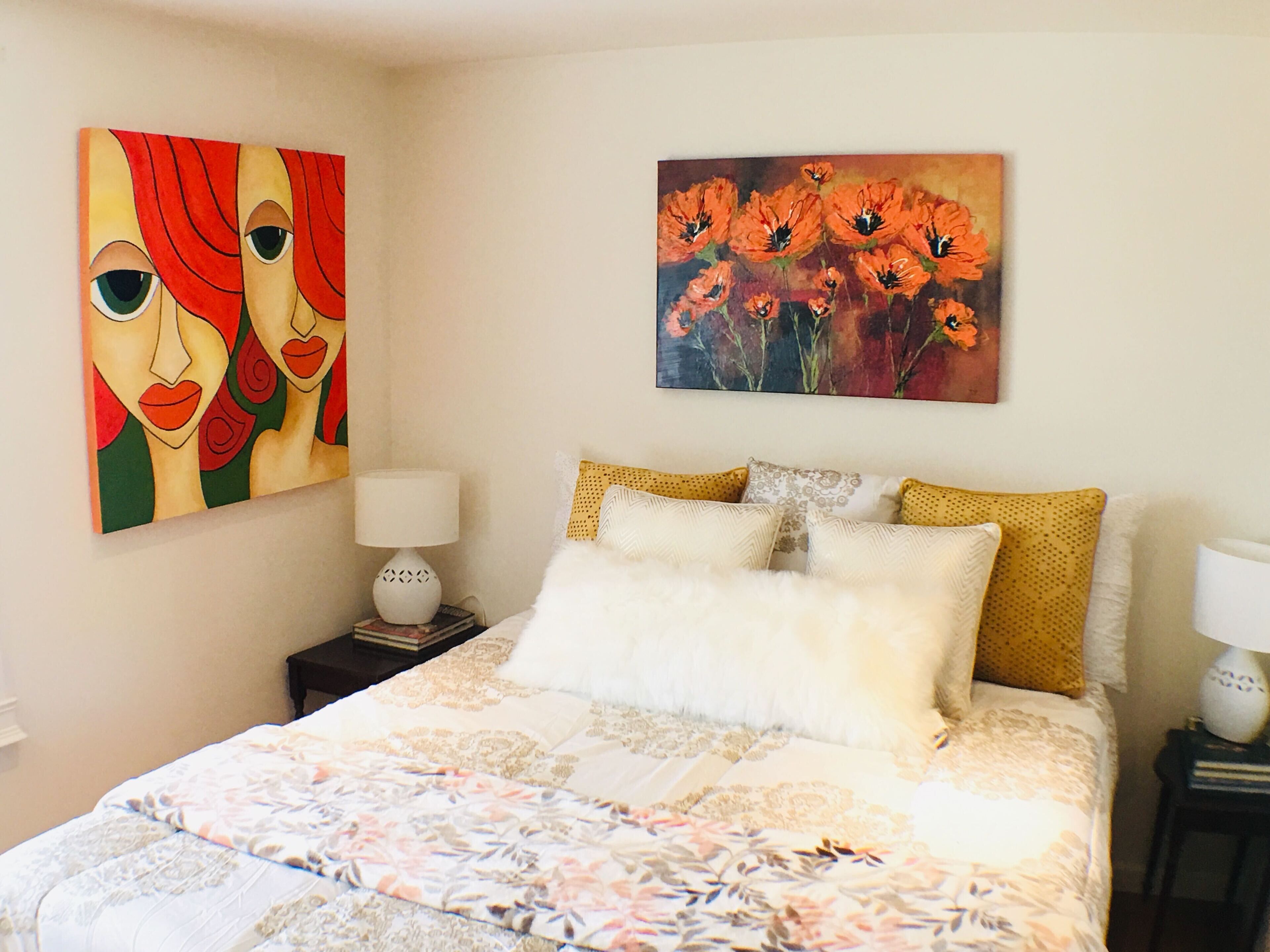 A bedroom with a large bed, two bedside tables, and two wall-mounted lamps. There are two paintings on the wall.