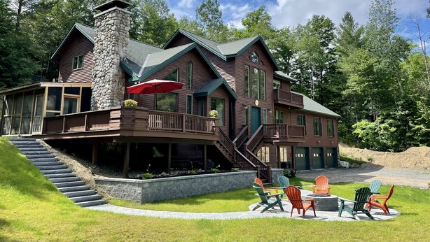 A two-story log cabin with a stone chimney, a covered porch, and a staircase leading to the upper level. - Cliff View Loj Home is in a Quiet Neighborhood Walking Distance to Mirror Lake. (Lake Placid)