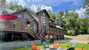 A two-story house with a stone foundation, a covered deck, and a fire pit area with Adirondack chairs. - Cliff View Loj Home is in a Quiet Neighborhood Walking Distance to Mirror Lake. (Lake Placid)