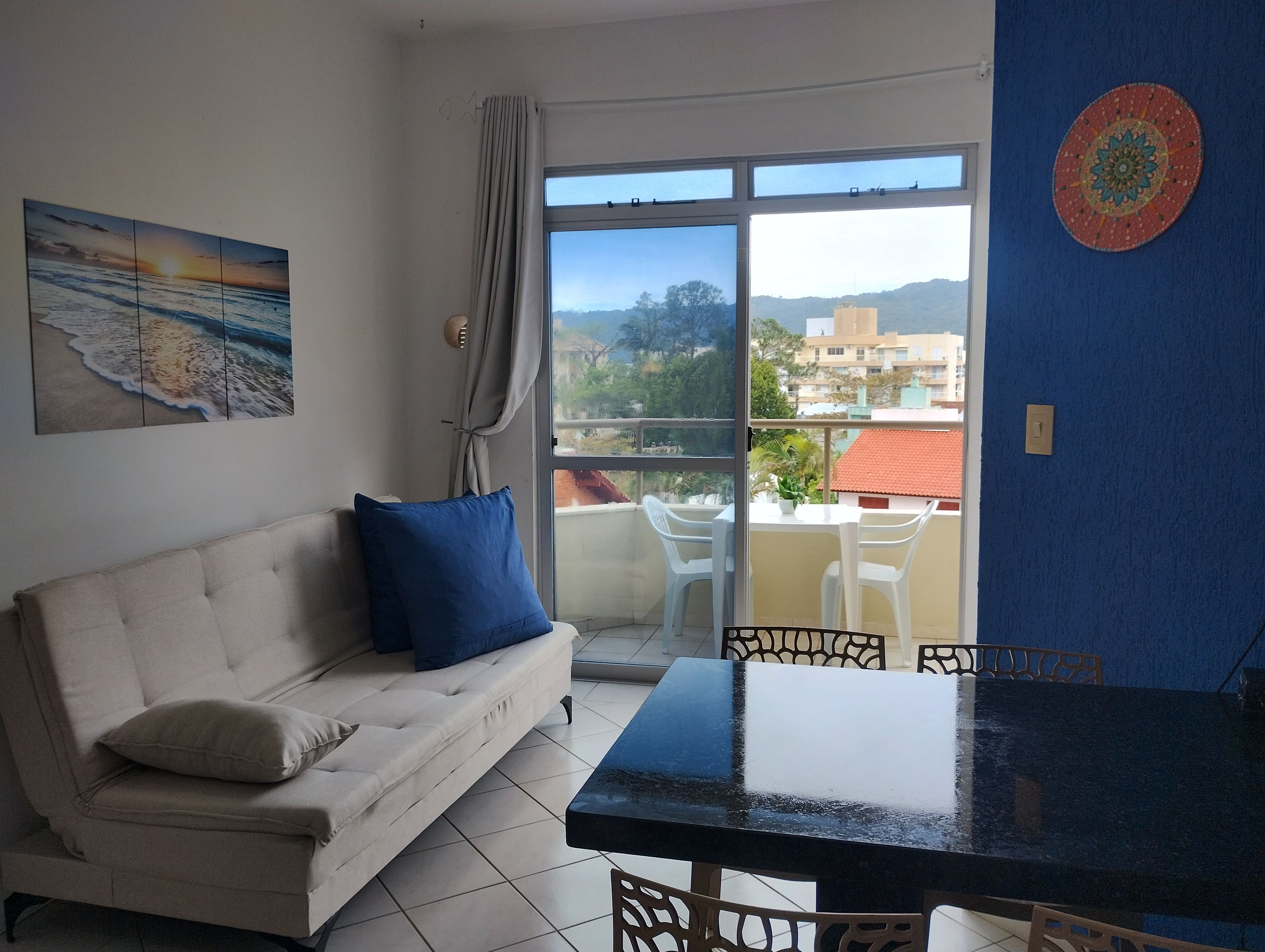 A living room with a sofa, a dining table, and chairs. There is a large window with a view of buildings and trees.