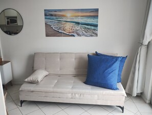 A beige sofa with a blue pillow and a white pillow, a wall-mounted picture of a beach scene, and a round mirror on the wall. - Apartment 50m from Cachoeira Beach - Balcony with BBQ, Air Conditioning and WiFi (Cachoeira do Bom Jesus)
