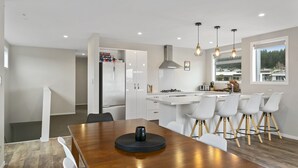 A modern kitchen with white cabinetry, a wooden dining table, and bar stools. There is a large window providing a view of the outdoors.