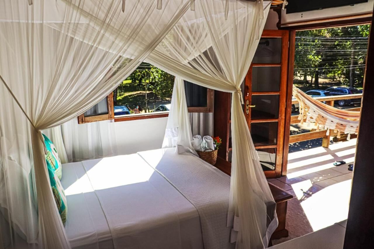 A room with a bed covered by sheer curtains, a wooden bench, a wicker basket with towels, and a view of a hammock outside.