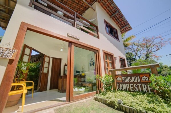 A two-story building with a reception area, a sign indicating 'Pousada Campestre', and a small outdoor seating area with yellow chairs. - Pousada Campestre (Porto Seguro)