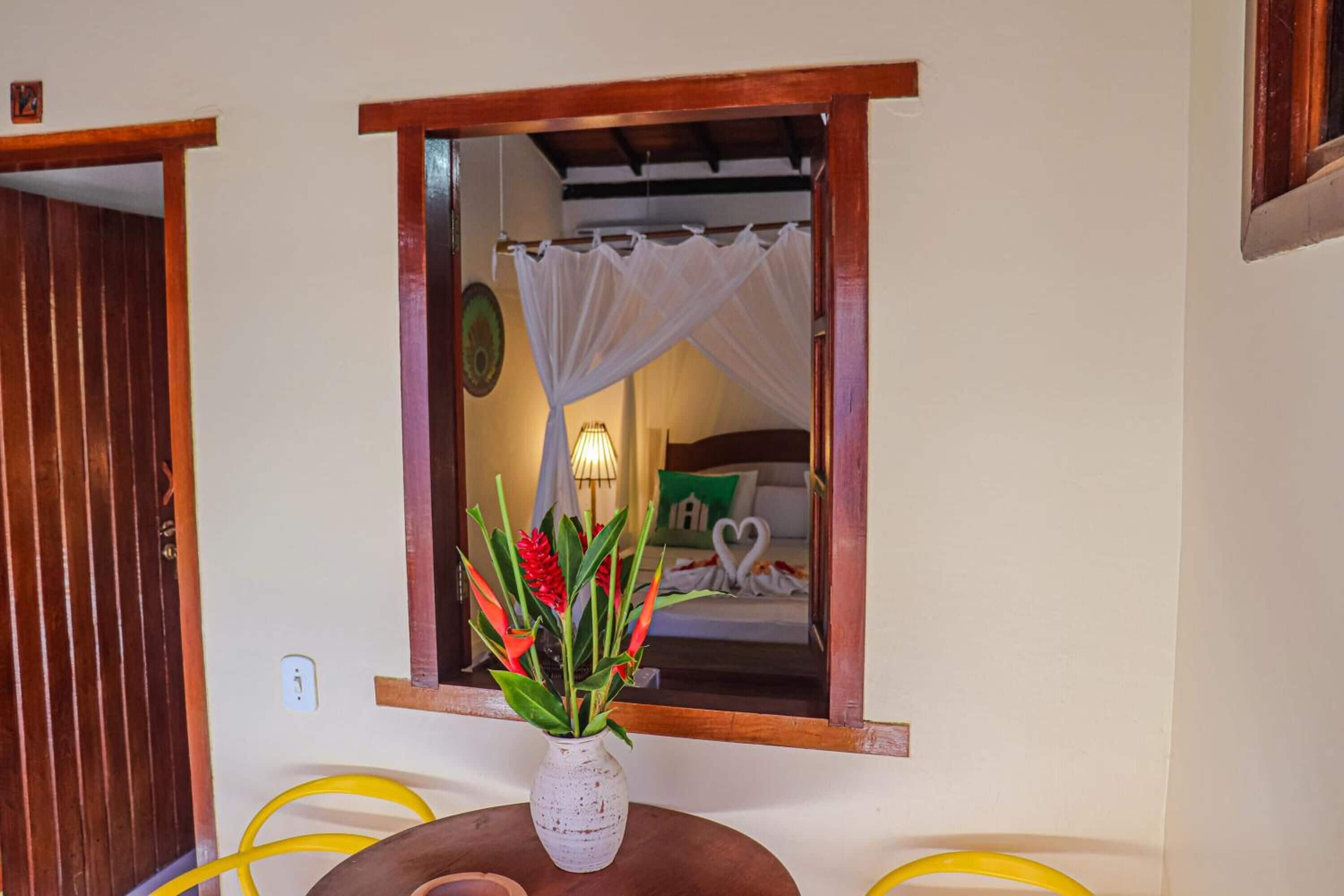 A room with a bed, a window with a white curtain, a vase with flowers, and a round table with a yellow object.