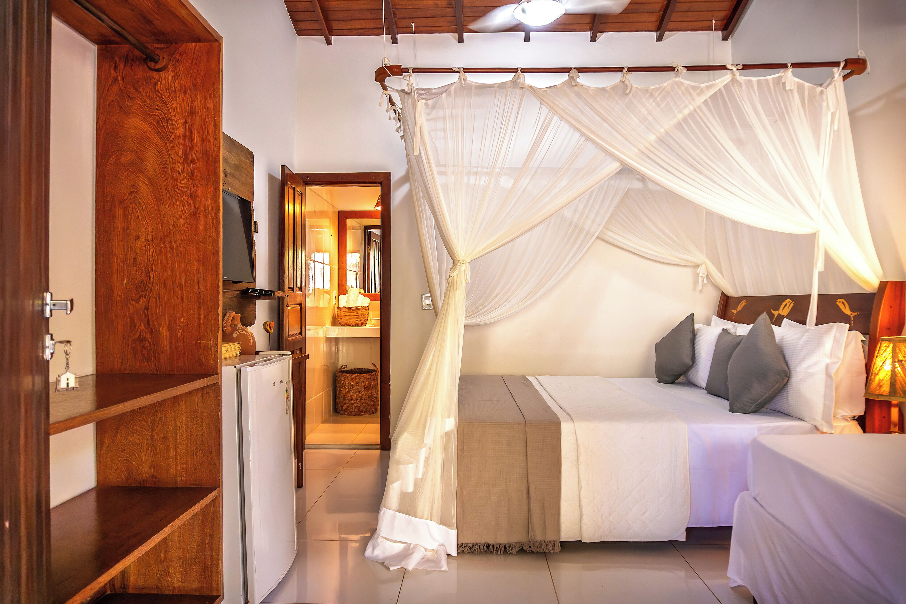 A bedroom with a canopy bed, a wooden wardrobe, a small refrigerator, and a bedside table with a lamp.