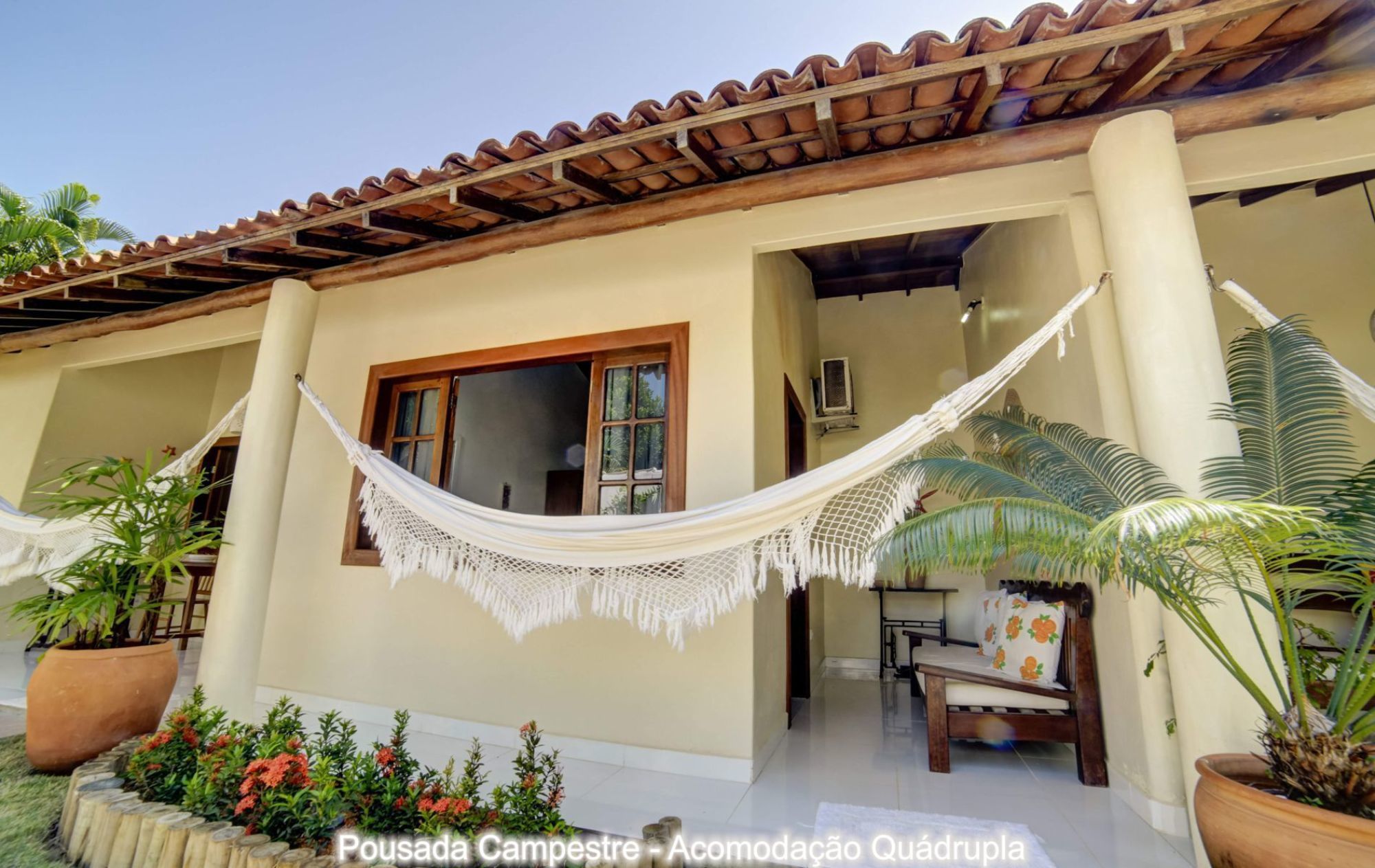 A hammock hanging between two columns in a covered outdoor area with a tiled roof, surrounded by potted plants and a wooden bench.