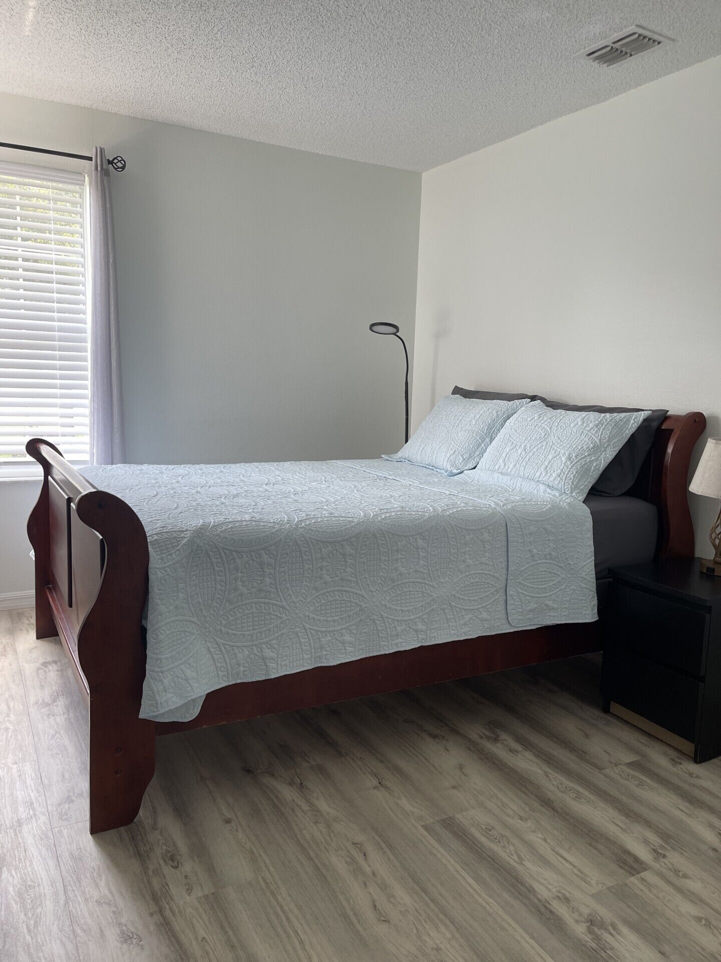 A single bed with a wooden frame and a light-colored quilt, a bedside table with a lamp, and a window with blinds.