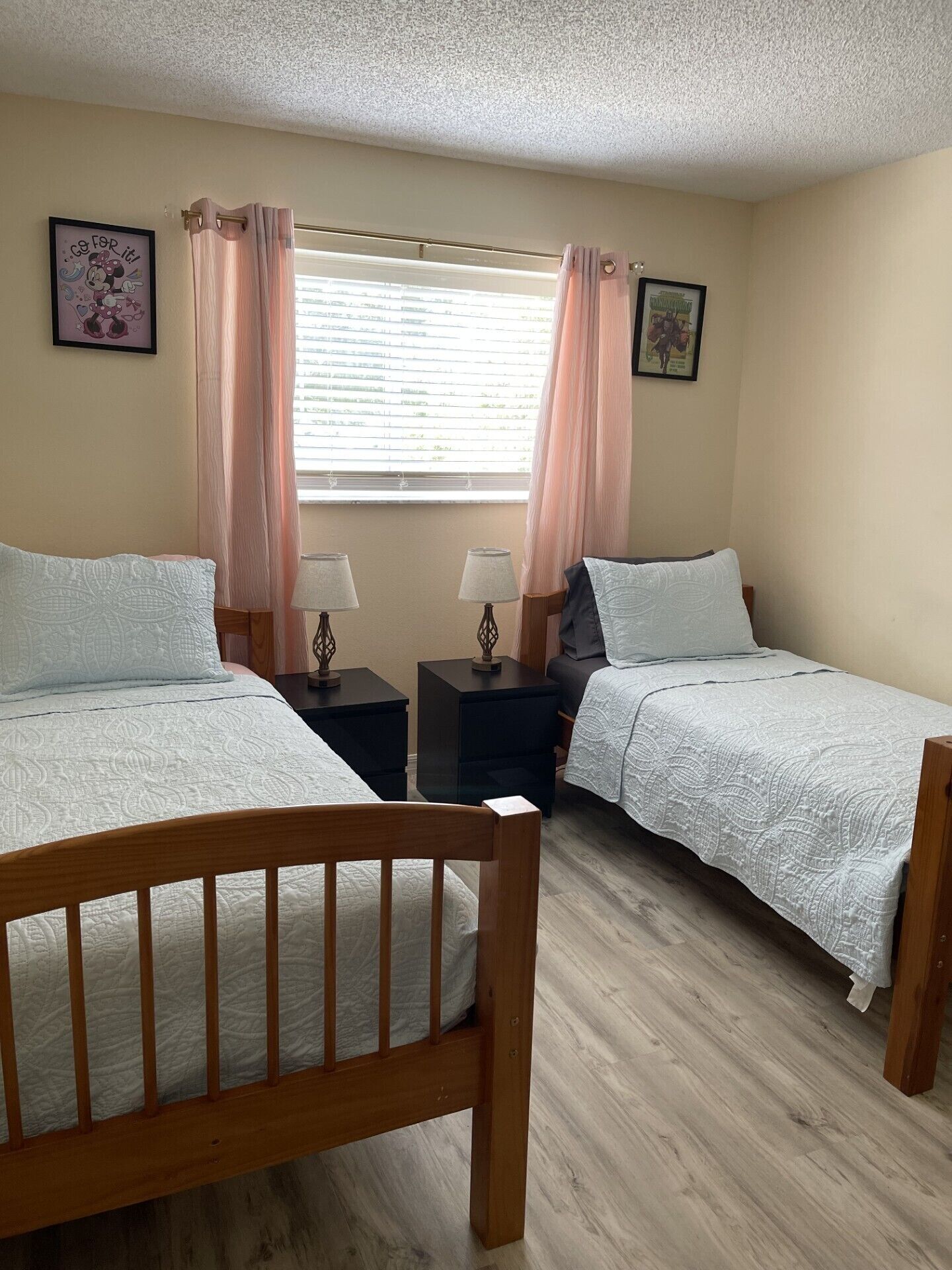 A bedroom with two beds, a window with curtains, bedside lamps, and two framed pictures on the wall.