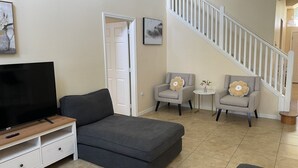 A living room with a grey sofa, a TV on a wooden stand, two armchairs, a small table with a vase, and a staircase. - 5 Bedroom Pool Home (Davenport)