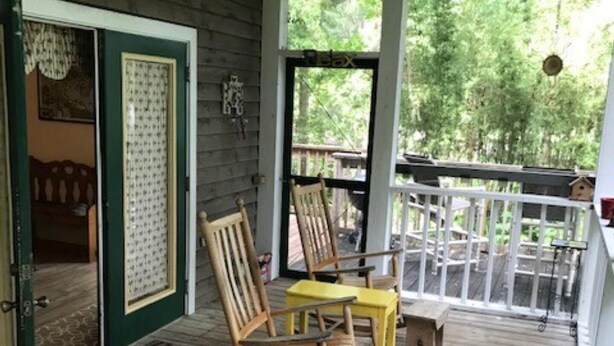 Eine hölzerne Veranda mit Schaukelstühlen und einem Tisch, eine Tür mit grünem Rahmen sowie ein Blick auf Bäume durch die Fenster.