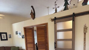 Ein gemütliches Wohnzimmer mit Holzboden, einem braunen Ledersofa, einem Holztisch und einem weißen Sessel. Eine Schiebetür mit rustikalem Design und ein wandhängendes Windspiel sind ebenfalls vorhanden.