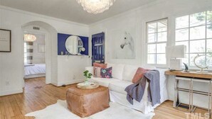 A spacious living room with a white couch, a wooden coffee table, and a desk with a globe. There is a large window providing natural light and a chandelier hanging from the ceiling. - Heart Of The City ❤️ Hot tub, ITB, NC State Cameron Village  (Raleigh)
