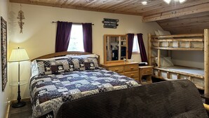 Ein gemütliches Zimmer mit Holzbalkendecke, einem Bett mit gemustertem Quilz, einem Nachttisch, einem Spiegel und einem Etagenbett.