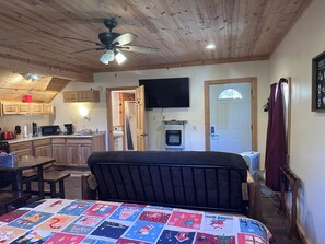 A bedroom with a bed, a ceiling fan, a television, and a view of the kitchen and bathroom. - Santa's Shack Studio - Near Pictured Rocks (Christmas)