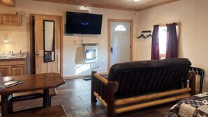 Ein Zimmer mit Holzboden, Deckenventilator, Flachbildfernseher, Bett, Schreibtisch mit Stuhl, Waschbecken und Spiegel.