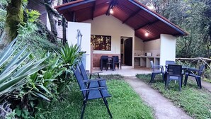 A covered outdoor seating area with chairs and a table, surrounded by lush greenery and a pathway leading to a building with a tiled floor and a window. - Bosques del Tibet (Santa Ana)