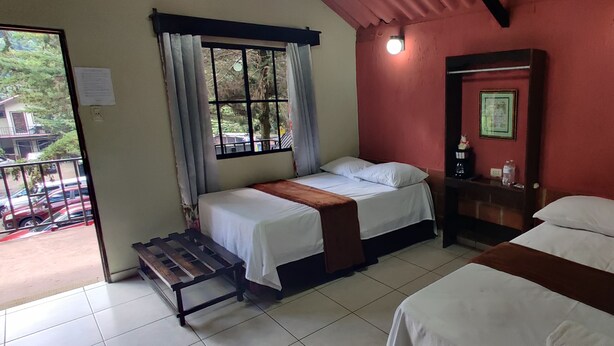 A room with two beds, a bench, a window with curtains, a mirror, and a wall-mounted shelf. - Bosques del Tibet (Santa Ana)