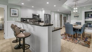 A modern kitchen with white cabinets, a granite countertop, and stainless steel appliances. There is a bar area with stools and a dining table with chairs. - Treasure Island 0805 (Panama City Beach)