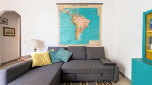 Un salon avec un canapé d’angle gris, une carte accrochée au mur, une lampe et une bibliothèque.