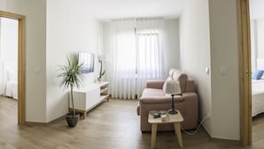 Una sala de estar moderna y minimalista con un sofá, una mesita de noche con una lámpara, una planta en maceta y un televisor sobre un mueble blanco.