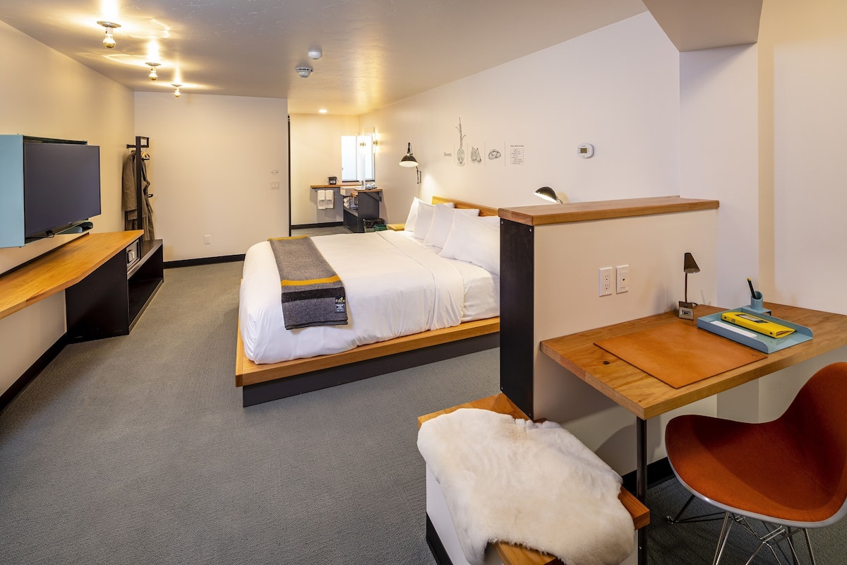 A modern hotel room with a bed, a desk, a chair, a TV, and a small kitchenette.