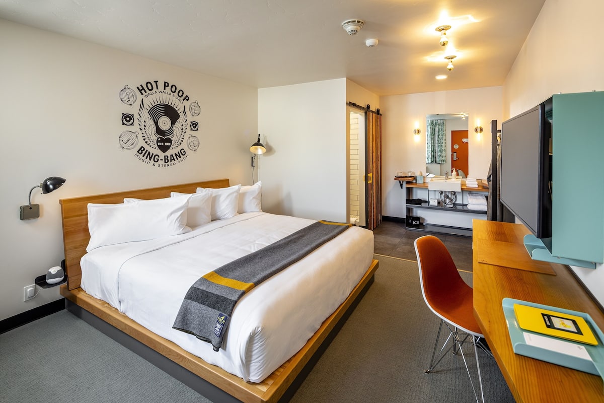 A hotel room with a bed, a desk, a chair, a TV, and a bathroom visible through an open door.