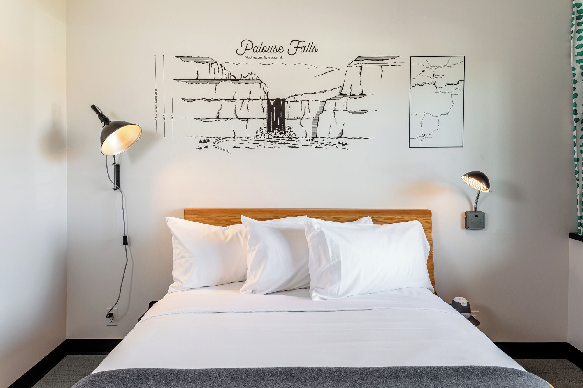 A hotel room with a bed, two wall-mounted lamps, and a wall art depicting Palouse Falls.