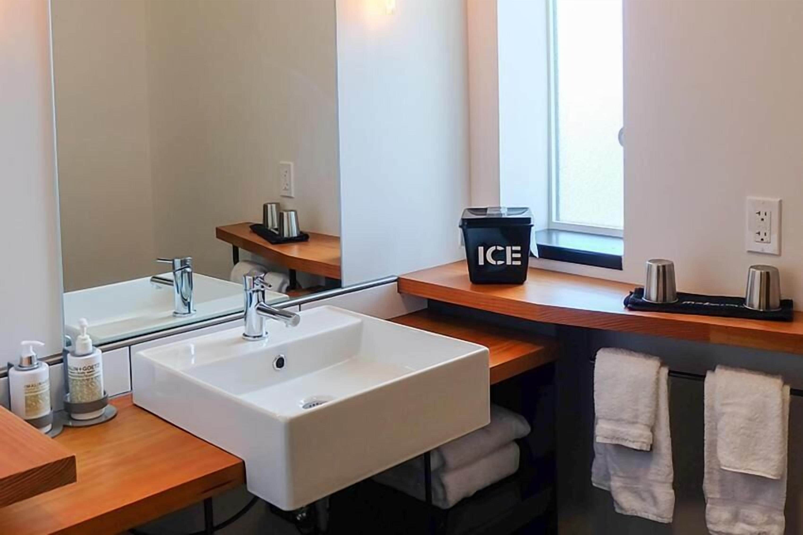 A modern bathroom with a white sink, mirror, and wooden countertop. There is a black trash bin labeled "ICE" and a window providing natural light.