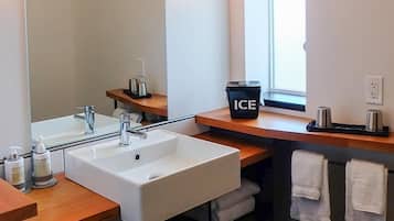 A modern bathroom with a white sink, mirror, and wooden countertop. There is a black trash bin labeled "ICE" and a window providing natural light.
