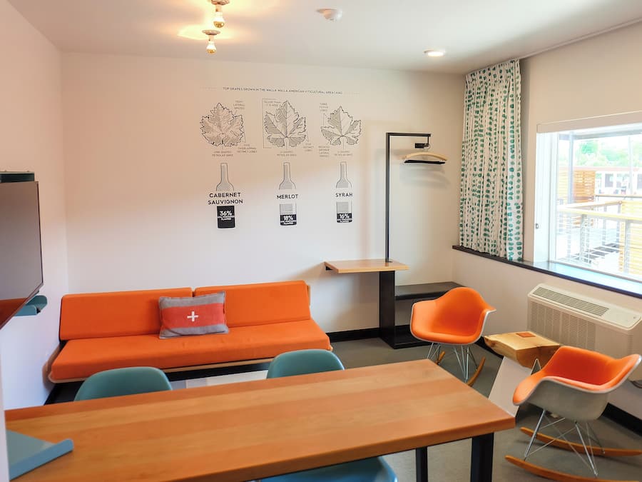 Una habitación moderna con un sofá naranja, dos mecedoras, una mesa de madera y un amplio ventanal con cortinas.