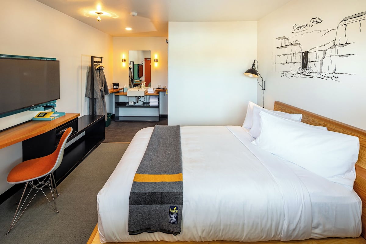A hotel room with a bed, a desk with a TV, a chair, a bathroom with a mirror, and a wall with a drawing.