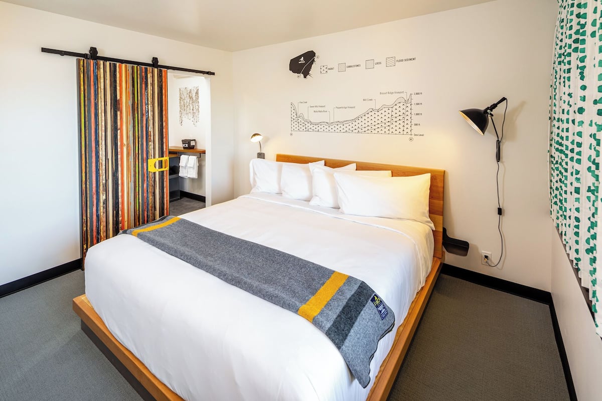 A hotel room with a large bed, a wooden headboard, a map on the wall, a desk with a chair, and a bathroom with a towel rack.
