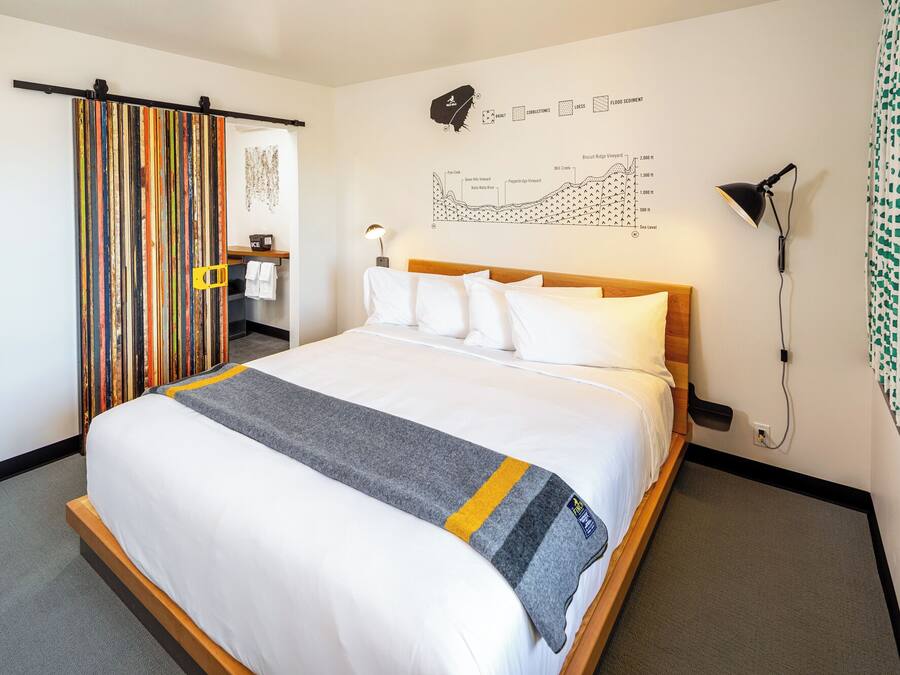 Una habitación de hotel con una cama grande, cabecero de madera, un mapa en la pared, un escritorio con silla, y un baño con perchero.