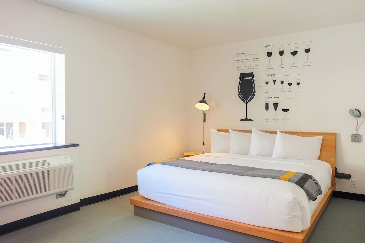 A hotel room with a bed, a wall-mounted light, a window, and a wall chart.